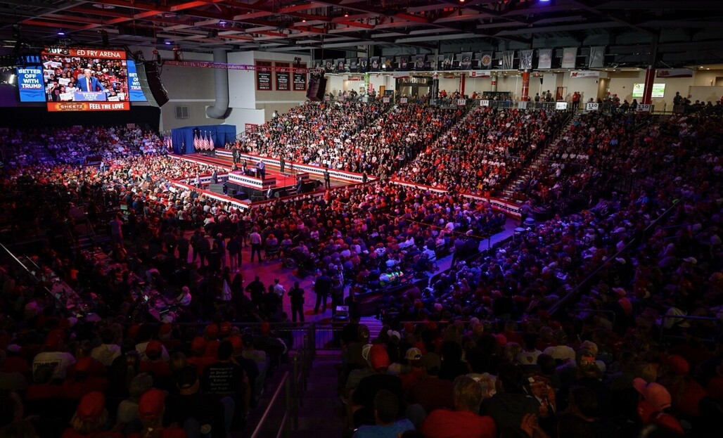 Packed indoor arena filled with a large audience attending a political rally, with a stage at the front adorned with American flags and banners. A speaker addresses the crowd, which is seated in tiered sections. The venue displays prominent signage including messages "Better Off With Trump" and "Feel The Love." Several overhead screens broadcast the event to the audience.
