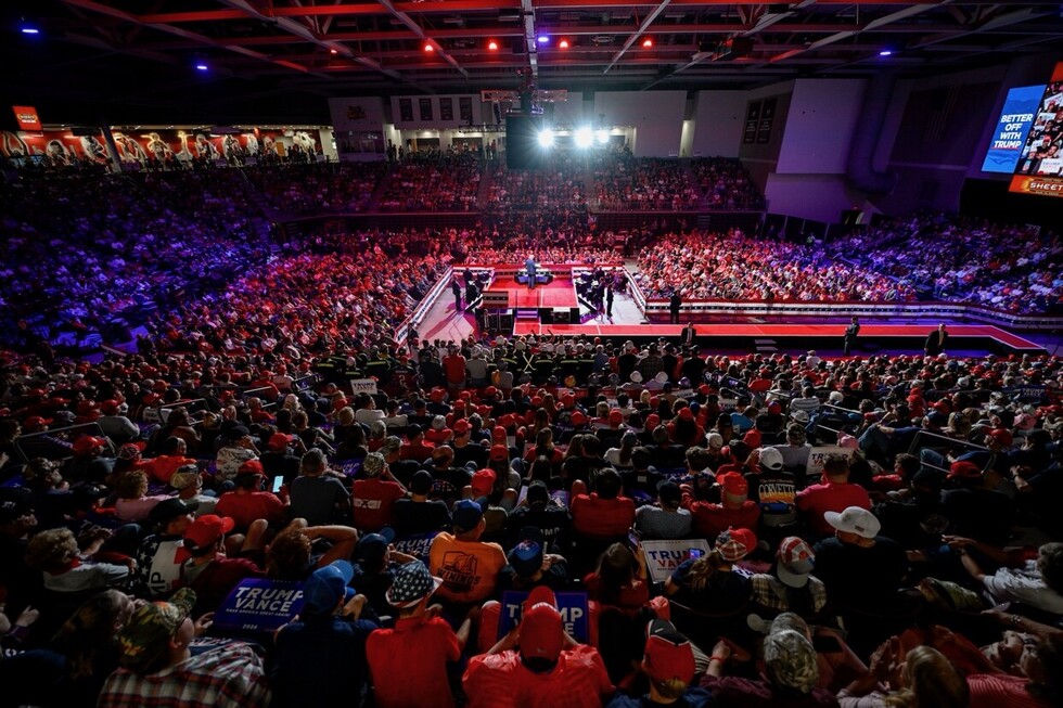 Large crowd at a political rally in an indoor arena, many people wearing red hats and holding signs, elevated stage with speaker, overhead lighting, and large screens displaying messages such as "BETTER OFF WITH TRUMP."