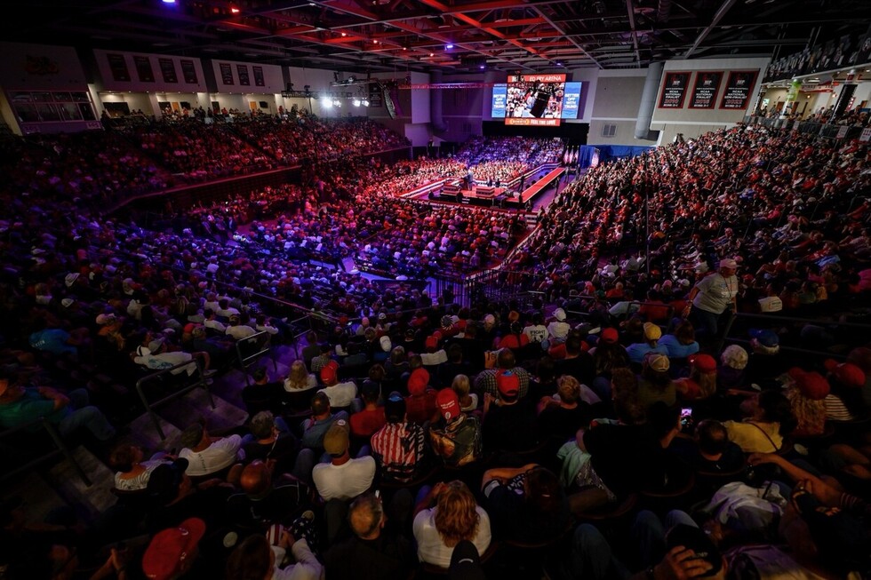 Large crowd attending an indoor event at an arena, with a central stage featuring a speaker. Stadium lighting casts red and blue hues over the audience.