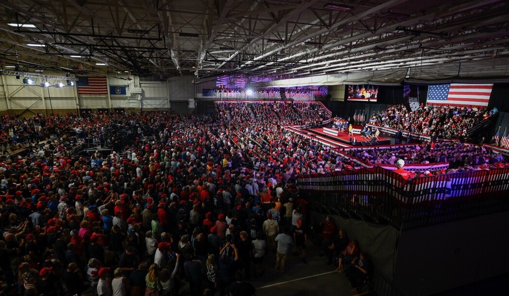 A large-scale indoor political rally with a crowded audience, many wearing red hats, facing a stage decorated with American flags. The venue is filled with people, and there is a notable presence of media equipment and personnel. A large American flag hangs prominently on the wall.