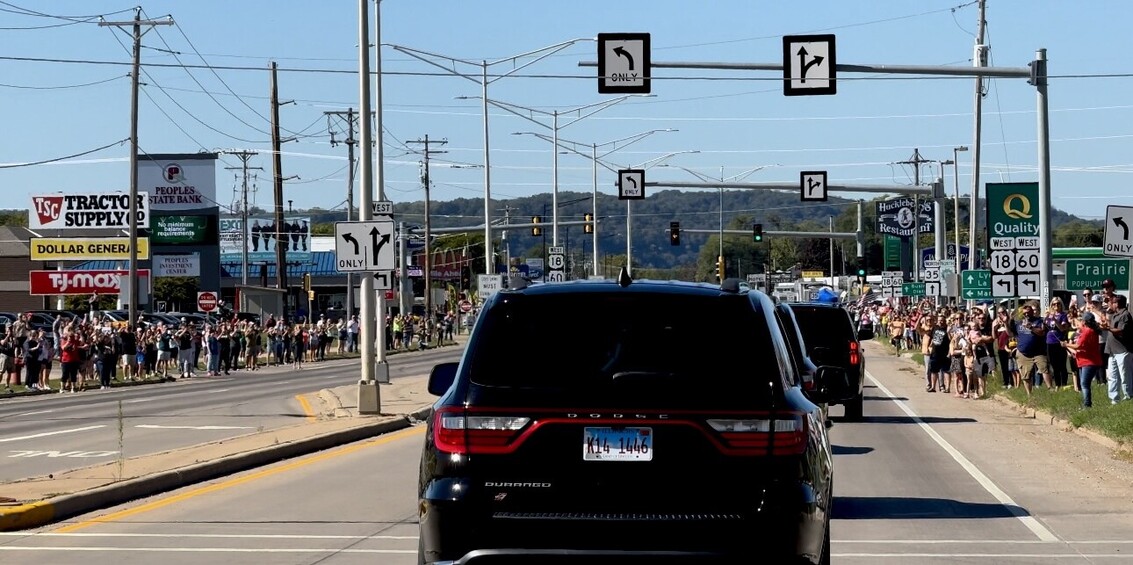 A line of black SUVs driving down a busy street with large crowds of people on both sidewalks, waving flags and banners. Numerous street signs and traffic lights overhead, commercial buildings and retail stores such as Tractor Supply, Dollar General, and TJ Maxx in the background. The scene suggests a procession or parade.