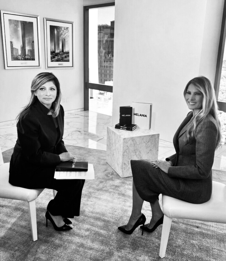 Two women seated on chairs in a modern, spacious room with large windows. One woman holds a book labeled "MELANIA" while the other has a similar book placed on a marble table between them. The walls feature framed architectural photographs, and both women wear professional attire and high heels.