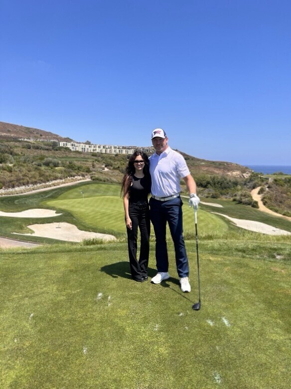 A man and woman standing on a golf course, with vibrant green grass and sand bunkers in the background; both look at the camera, with the man holding a golf club and wearing a white cap and glove, while the woman is in a black outfit. Rolling hills and modern buildings are visible in the distance under a clear blue sky.