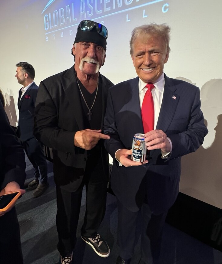 Two men stand together, one holding a can labeled "Real American" with an eagle design. They are in a well-lit room with a "Global Ascension Studios LLC" sign in the background. The man on the left is wearing a black bandana, sunglasses, a black jacket, and a cross necklace, pointing at the can. The man on the right is wearing a navy suit with a red tie and smiling. Other people are visible in the background.