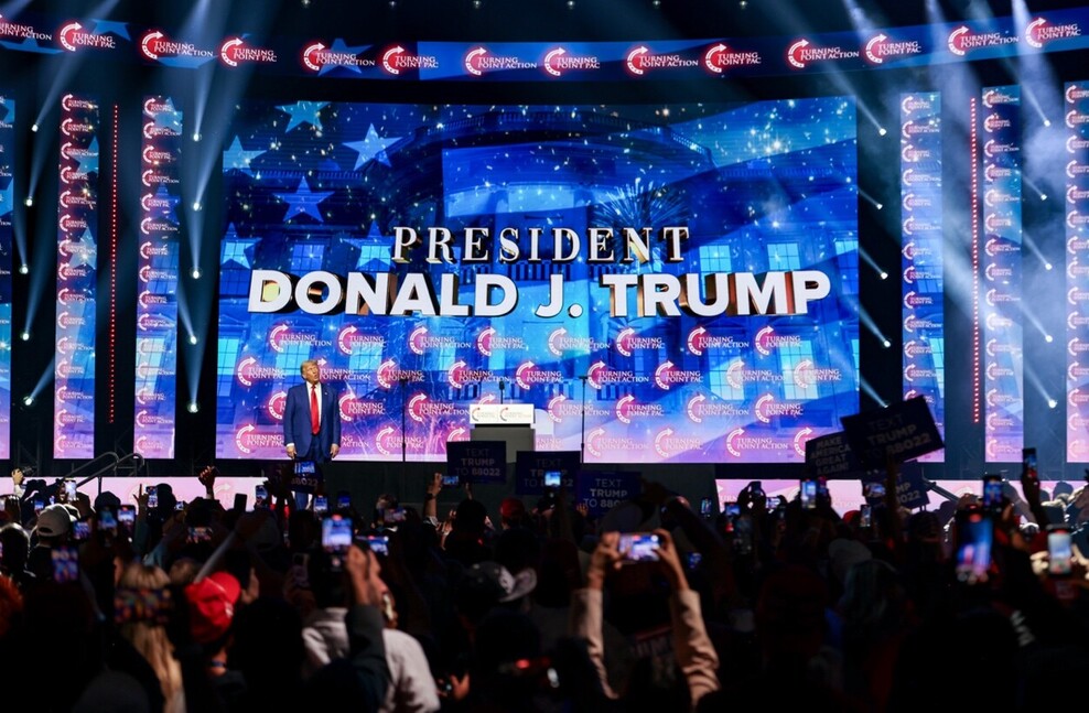 Donald J. Trump on stage at a Turning Point Action event, surrounded by large screens displaying his name with a crowd taking photos and holding signs.