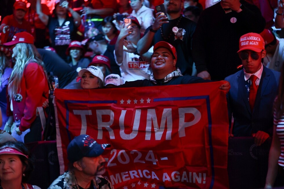 Supporters at a rally hold a red "Trump 2024 - Make America Great Again" banner. Attendees wear red hats and patriotic attire, capturing the event's enthusiastic and energetic atmosphere.