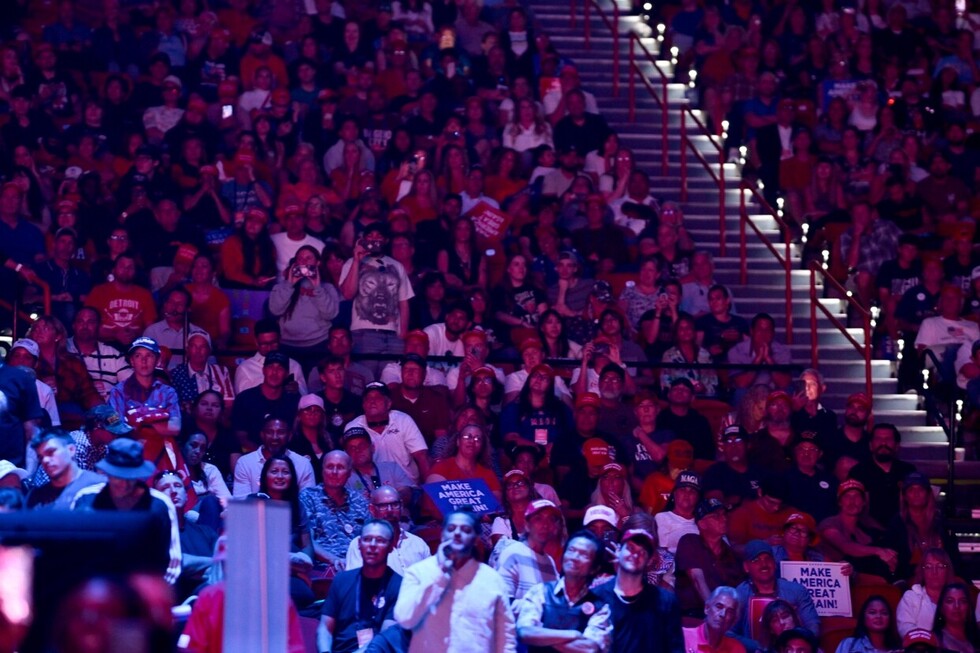 A large crowd of people seated in an arena, many wearing red hats and holding signs with "Make America Great Again" slogans. The atmosphere is lively, with attendees taking photos and engaging with the event.