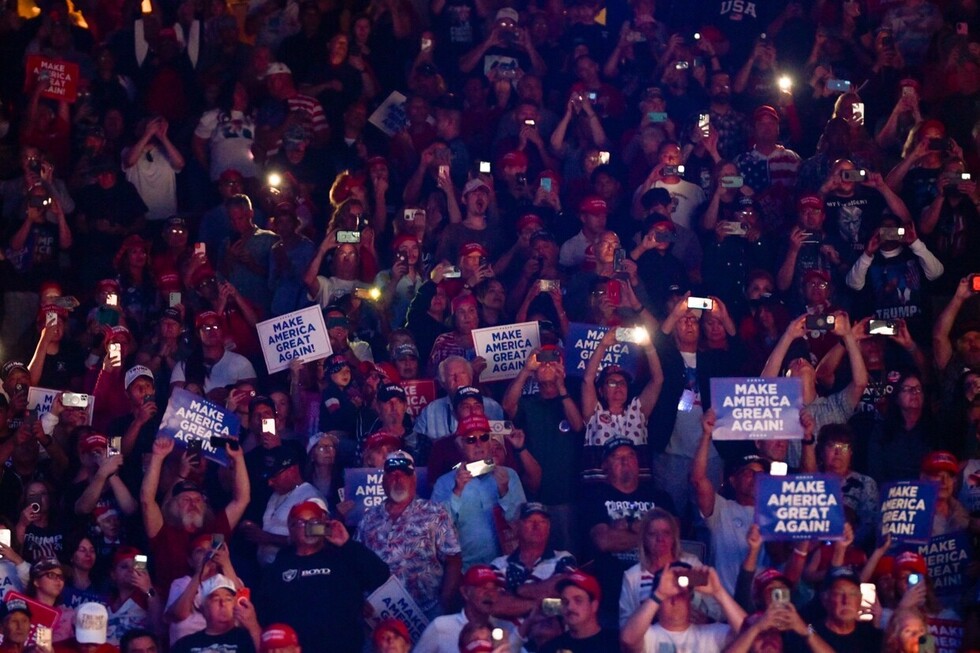 A crowd of people at a rally holding "Make America Great Again" signs and wearing red hats, many capturing the event with their phones.