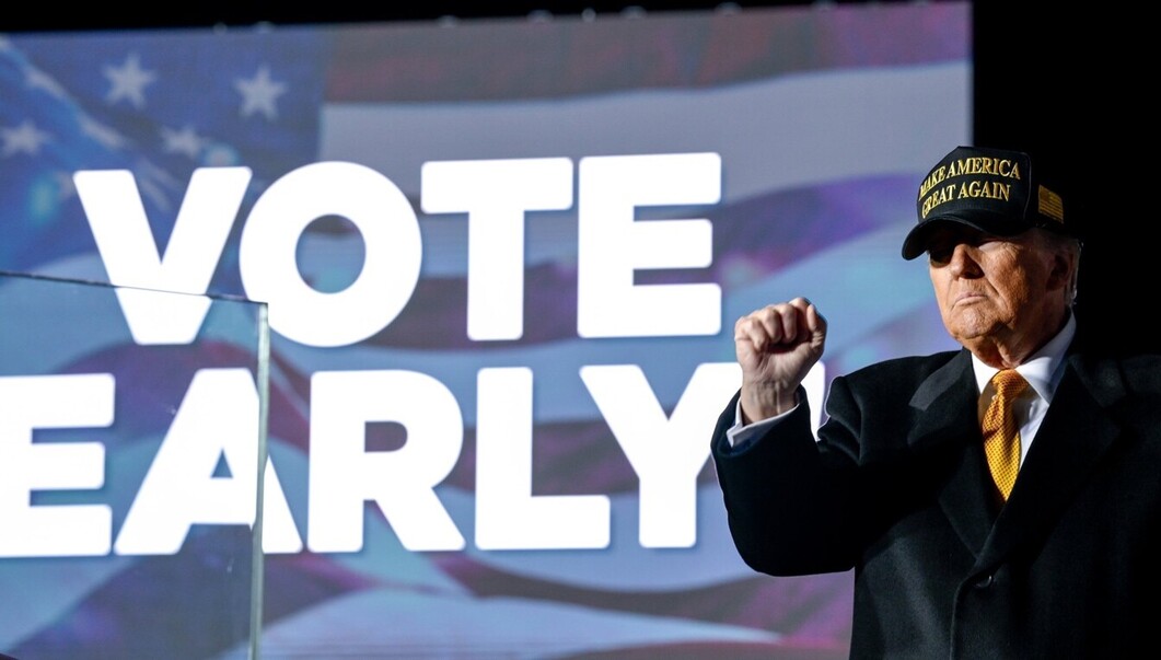 Man wearing a "Make America Great Again" cap, raising his fist, standing in front of a sign that reads "Vote Early" with an American flag background.