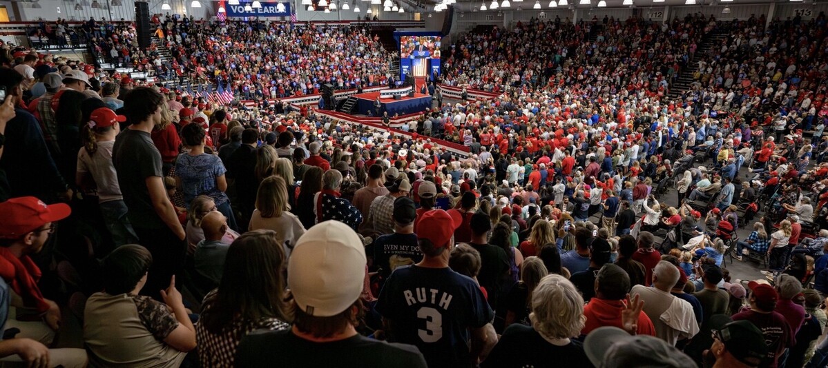 Large crowd attending a political rally in an indoor arena, with audience members wearing red hats and shirts. A stage is set up in the center with political signs and a speaker addressing the attendees, while American flags are displayed around the venue. The atmosphere is energetic and packed with spectators observing the event.