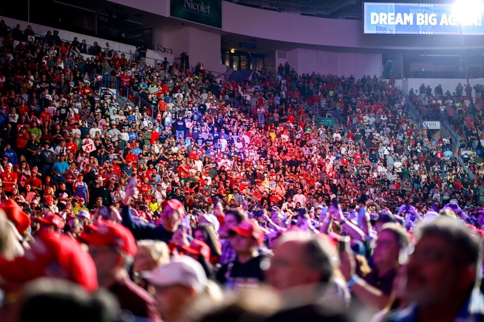 Large crowd at an indoor arena, with many people wearing red hats and shirts; a banner above reads "Dream Big Again."