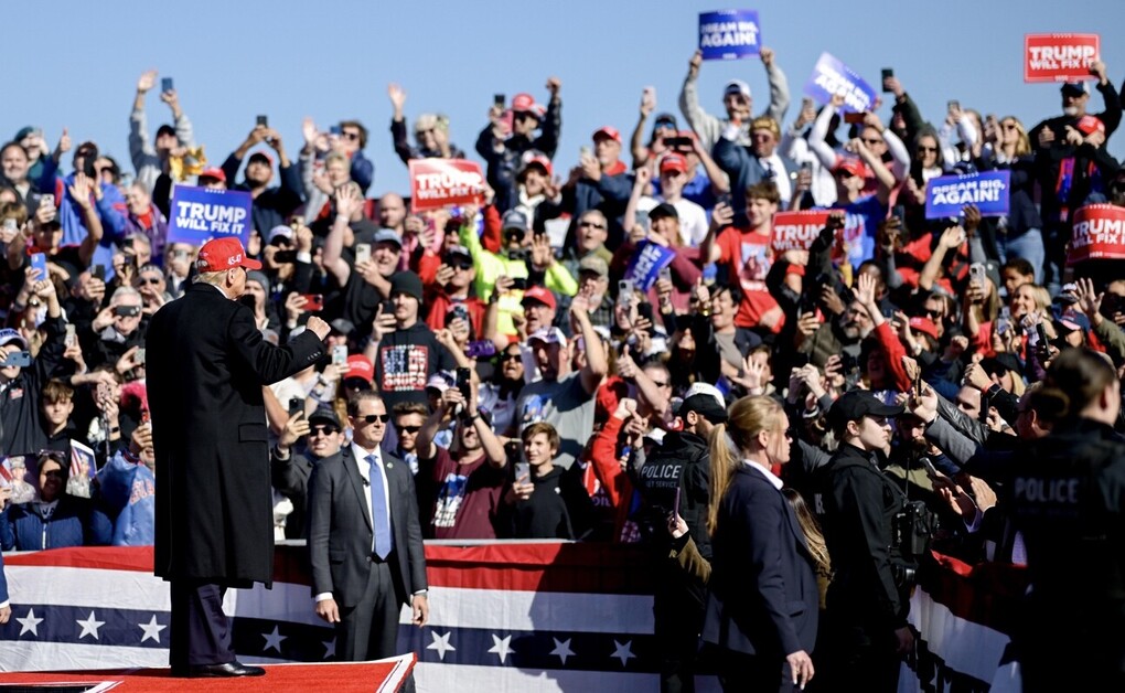 Large crowd at a political rally with people holding signs and wearing red hats, a prominent figure standing on stage addressing the audience, surrounded by security personnel. Banners and flags displaying political slogans.