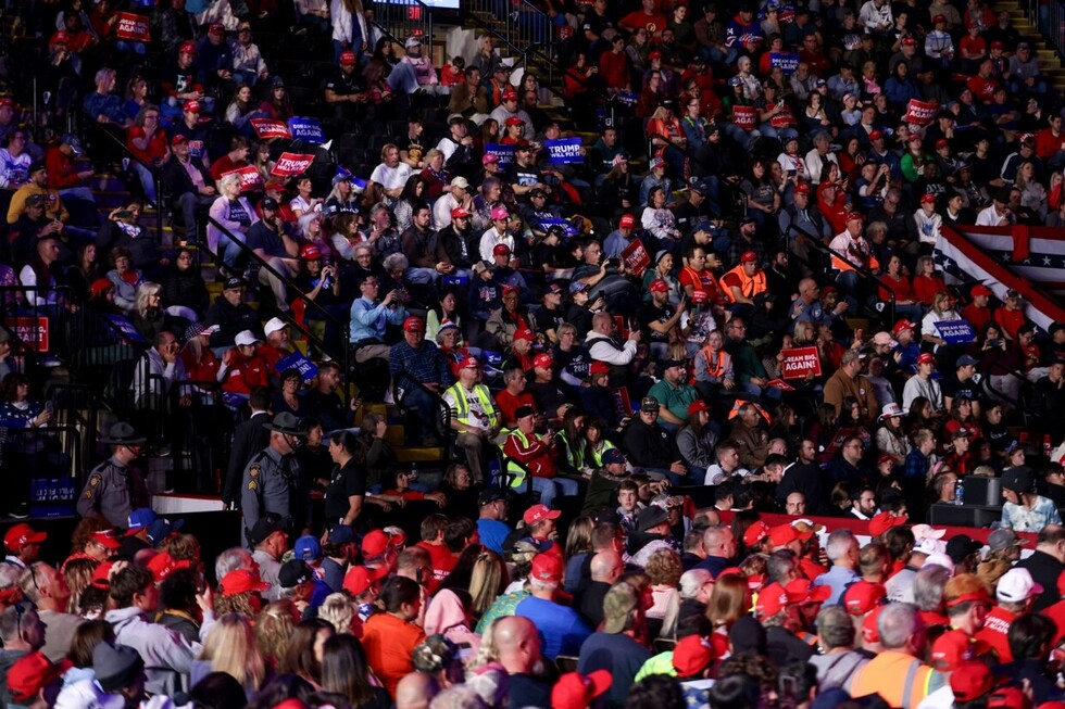 A crowded stadium filled with people wearing red hats and holding signs with slogans. Many attendees are dressed in patriotic colors, with event staff and security personnel visible. The seating area is packed, indicating a large gathering or rally.