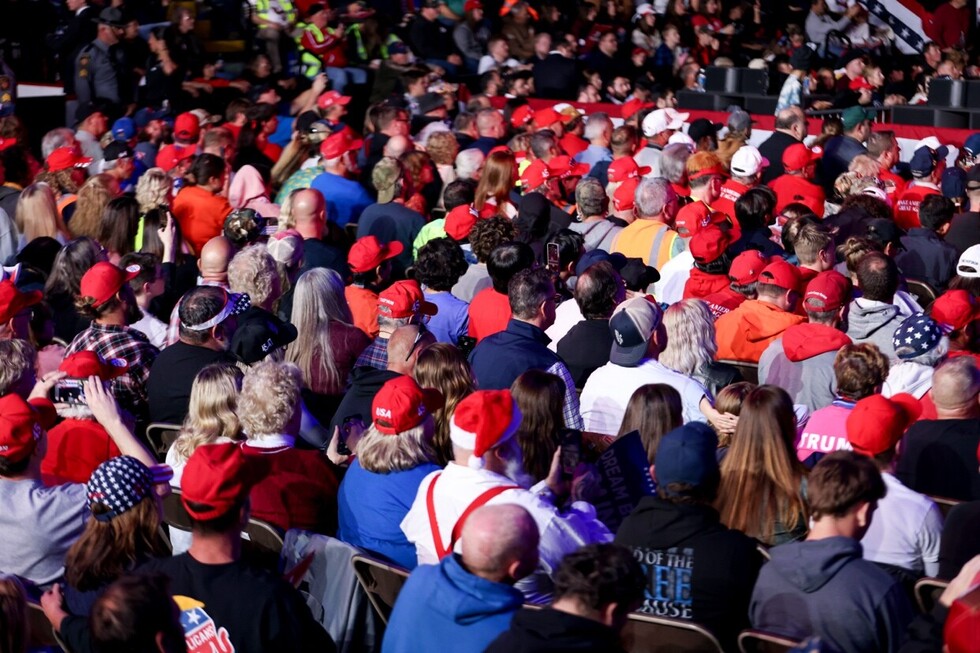 Large crowd wearing red hats and patriotic attire, gathered at a political rally, seated closely together, with people capturing the event on their phones.