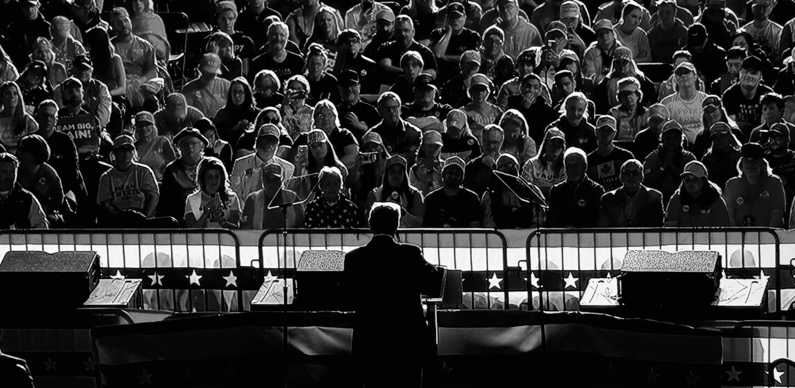 Black and white image of a speaker addressing a large crowd, with the audience seated behind a barrier decorated with stars and stripes. The speaker is silhouetted against the well-lit crowd, creating a dramatic contrast. Spectators are varied, including many wearing hats and holding cameras.