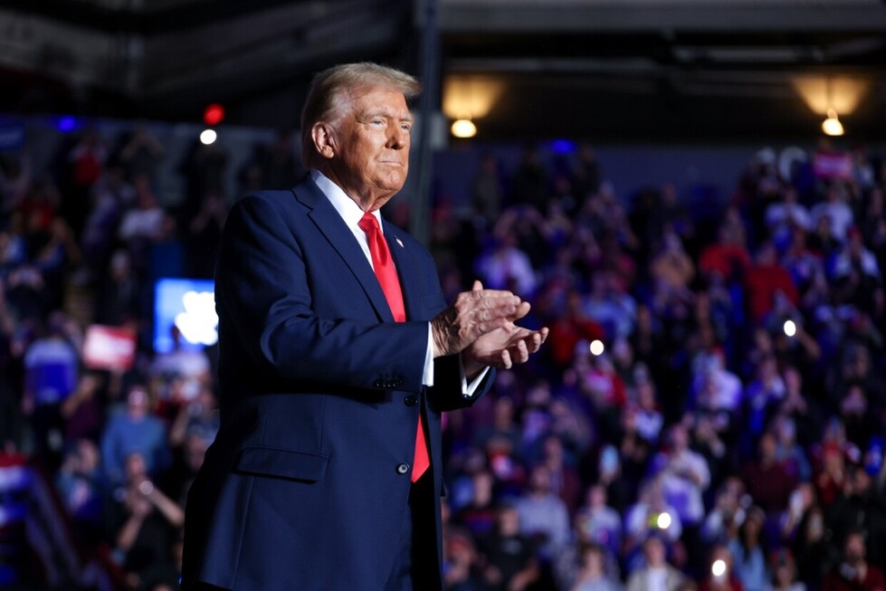 A man in a suit with a red tie stands on a stage, clapping his hands, with a blurred crowd in the background, some holding signs, in a large indoor venue.