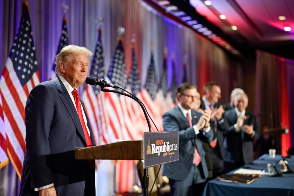 Politician speaking at a podium with a sign reading "House Republicans 118th Congress." Surrounded by clapping individuals in suits, in a room decorated with American flags.