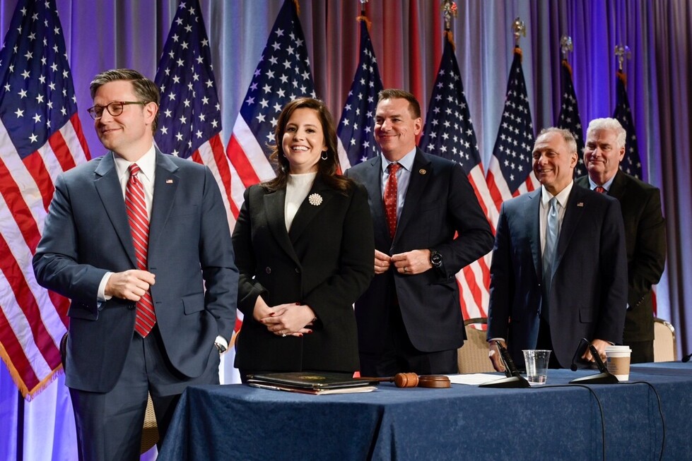 Group of five individuals in formal attire standing in front of multiple American flags on a stage, with a table holding documents and a gavel in the foreground.