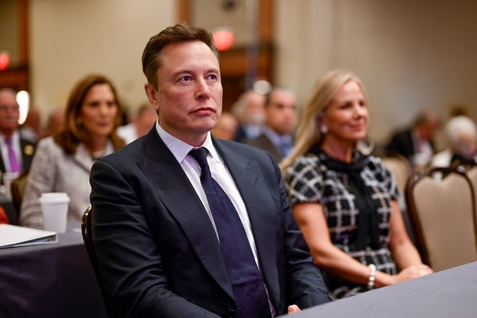 A man in a suit and tie sits attentively in a conference room, surrounded by other attendees. The setting is formal, with a blurred background indicating a large gathering or event atmosphere.