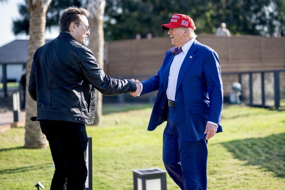 Two people shaking hands outdoors, one wearing a black leather jacket and sunglasses, the other in a blue suit with a red "Make America Great Again" hat. They stand on a grass field with trees and a fence in the background.