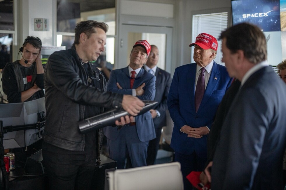 Group of men in formal attire inside a room, one holding a small rocket model; two wear red hats with a slogan. SpaceX logo visible on a screen.