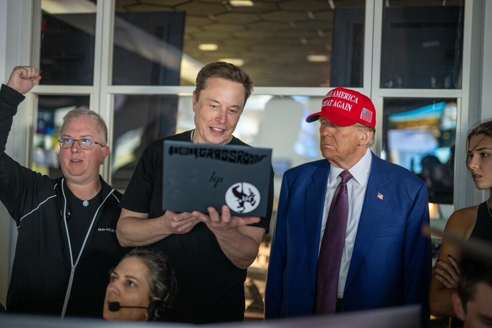 A group of people is gathered around a person holding a laptop, with one person wearing a red cap that has white text on it. They appear to be having a discussion in an indoor setting.