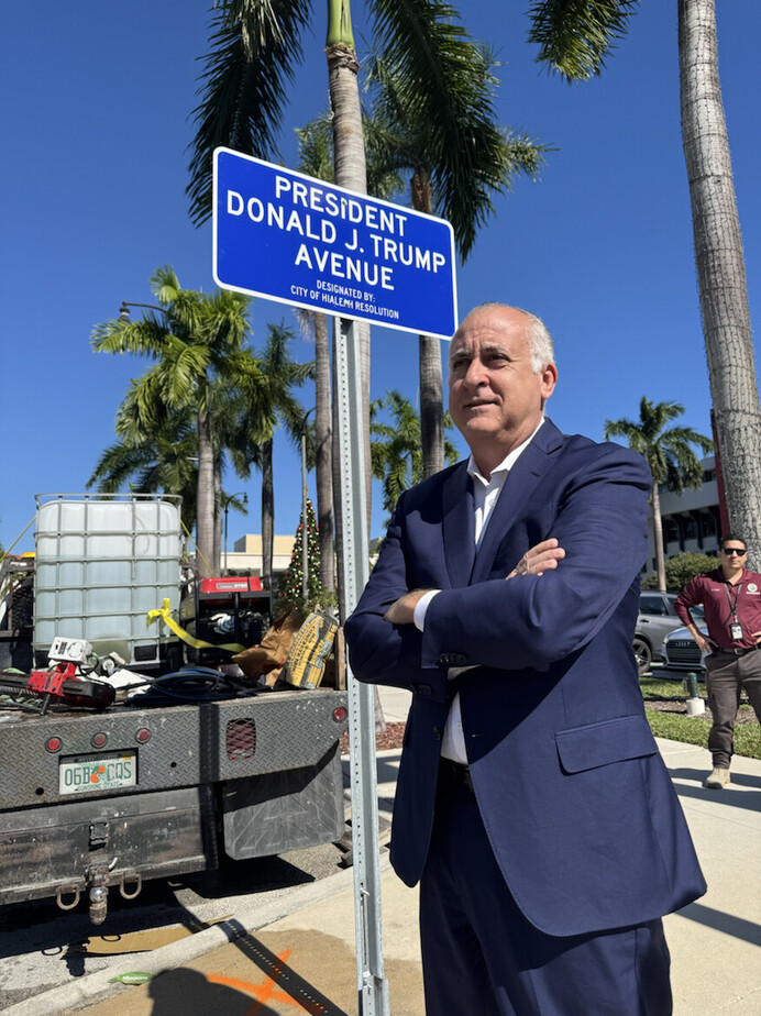 Man in a blue suit standing with arms crossed in front of a "President Donald J. Trump Avenue" street sign, surrounded by palm trees and a clear blue sky, with work equipment visible on a vehicle in the background.