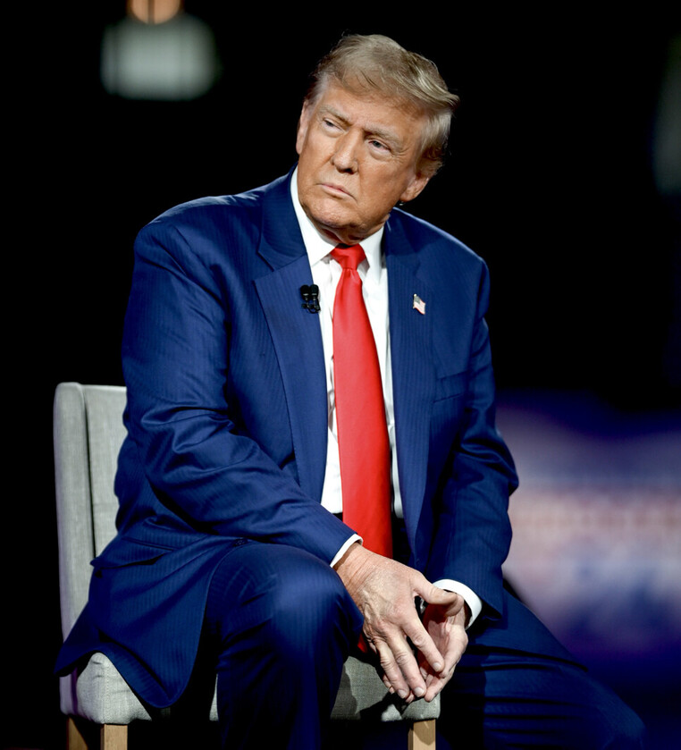 Man in a blue suit with a red tie sits on a chair on stage, looking to the side, with a microphone clipped to his jacket.