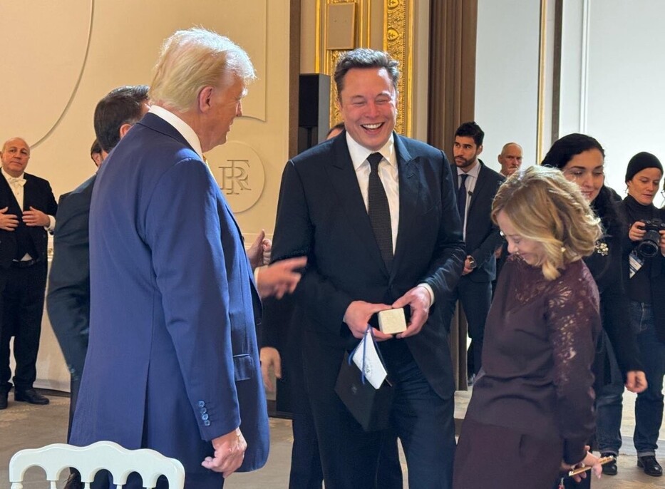 A group of people in formal attire are engaged in conversation at an indoor event. One individual is laughing and holding an item that appears to be a small book or box. The setting is ornate, with elegant decor and other attendees in the background, including some with cameras.