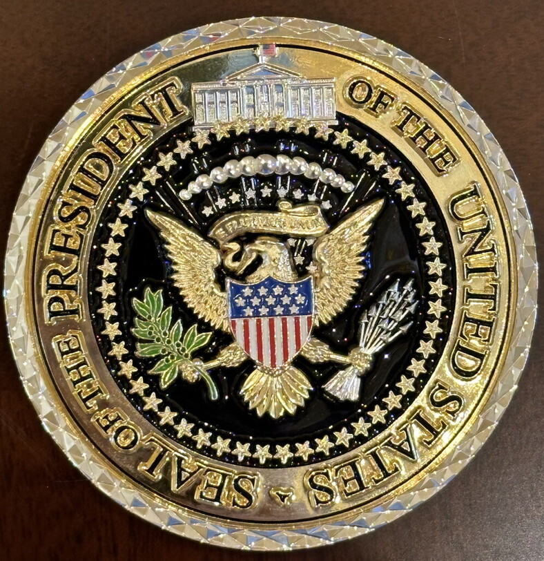 Presidential seal of the United States featuring a bald eagle holding an olive branch and arrows, with a shield on its chest. The design is enclosed within a gold border and the text "Seal of the President of the United States" surrounds the emblem.