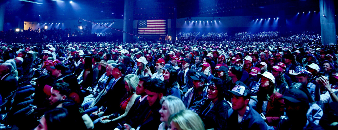 Large crowd wearing red and white hats in an indoor venue, with an American flag displayed in the background, illuminated by blue lighting.