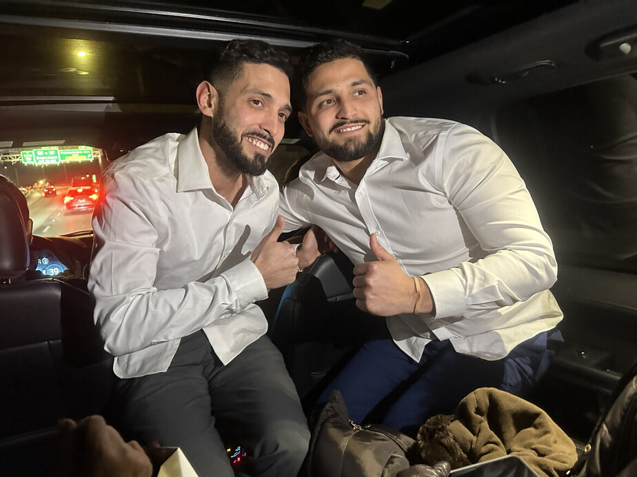 Two men sitting in the backseat of a car, both wearing white shirts, smiling and giving thumbs-up gestures. They are illuminated by the car's interior light, with traffic visible through the rear window.