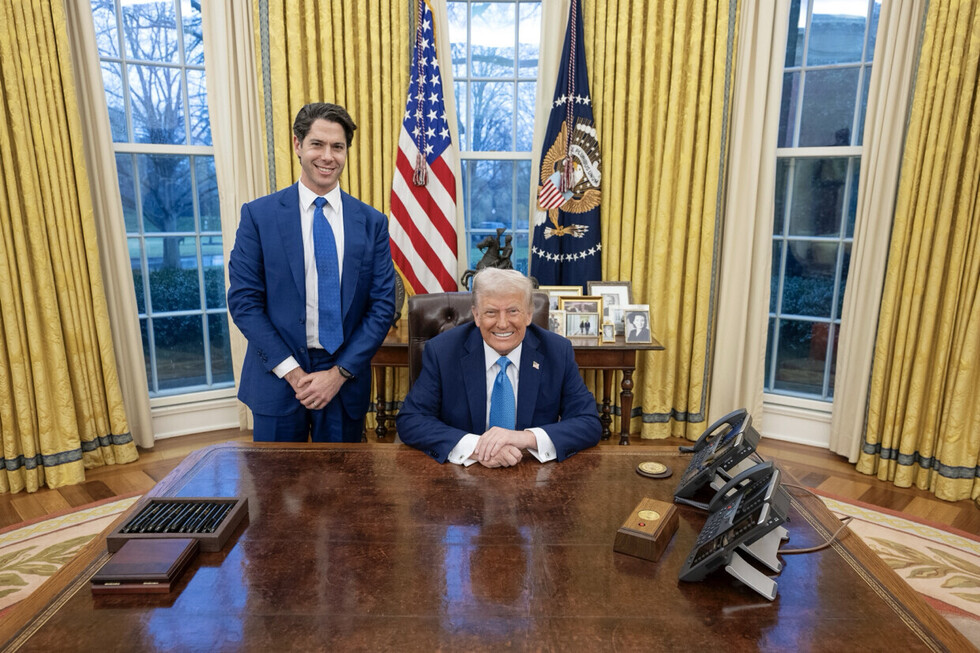 Two men in suits inside an office with yellow curtains, standing in front of a desk with telephones, photographs, and flags in the background. One is sitting; the other is standing.