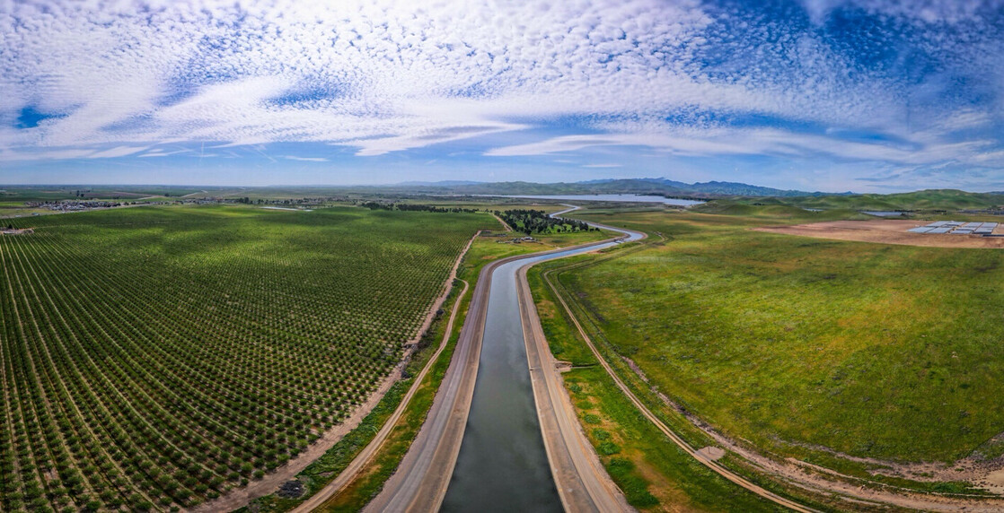 Aerial view of a winding canal running through expansive green fields and orchards under a blue sky filled with scattered clouds. The landscape includes lush agricultural land and distant rolling hills, creating a picturesque rural scene.