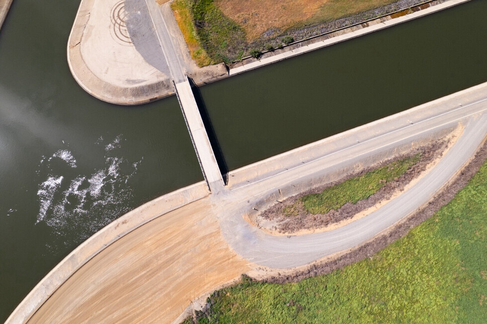 Aerial view of a canal with a concrete bridge crossing over it. The waterway features calm, dark green water, with a section displaying some white water turbulence. Adjacent to the canal, there are areas of grass and bare earth, with a curving road running parallel to the waterway.
