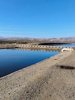 A tranquil water reservoir with a clear blue sky, surrounded by a gravel path and distant mountains. Reflections of the sky and landscape are visible on the water's surface.