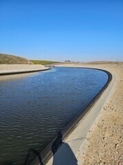 A curved man-made canal under a clear blue sky, with water flowing between concrete embankments and dry, dusty land on either side.