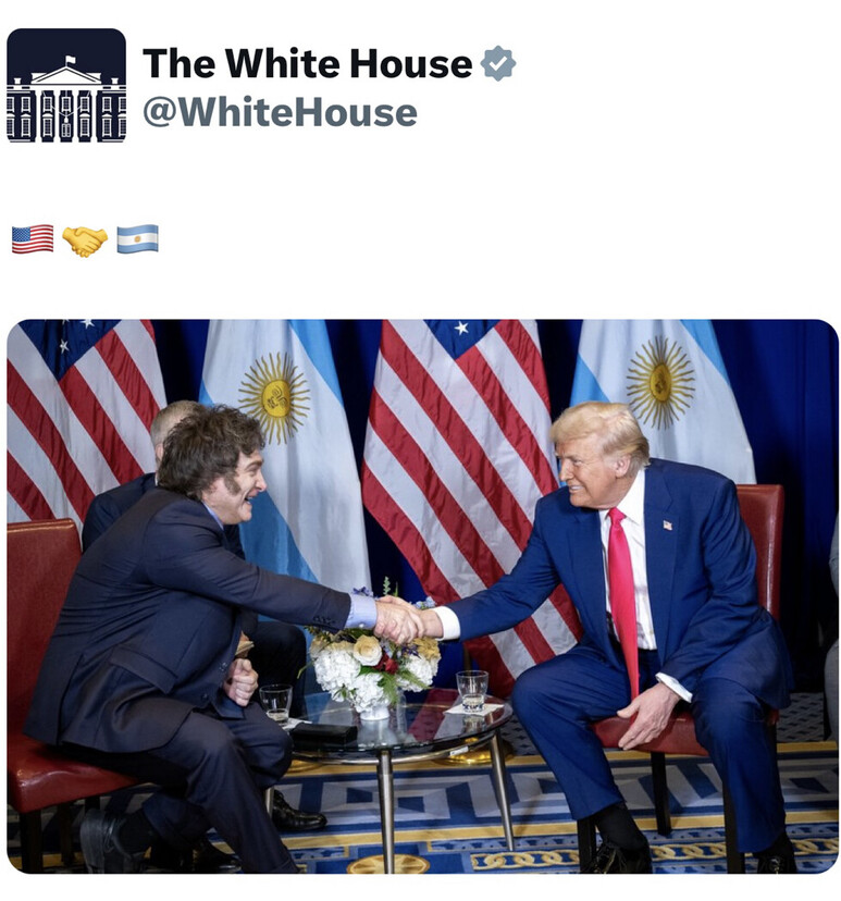 Two men in suits shaking hands in a formal setting with American and Argentinian flags in the background. One man has light hair, wearing a red tie and blue suit, seated on the right. The other man has dark hair and a dark suit, seated on the left. A small round table with flowers and glasses is positioned between them.