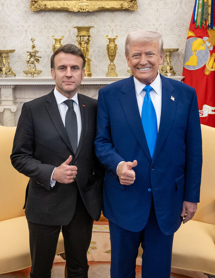 Two men in suits standing side by side with their thumbs up, in an elegantly decorated room with golden ornaments and a flag in the background.
