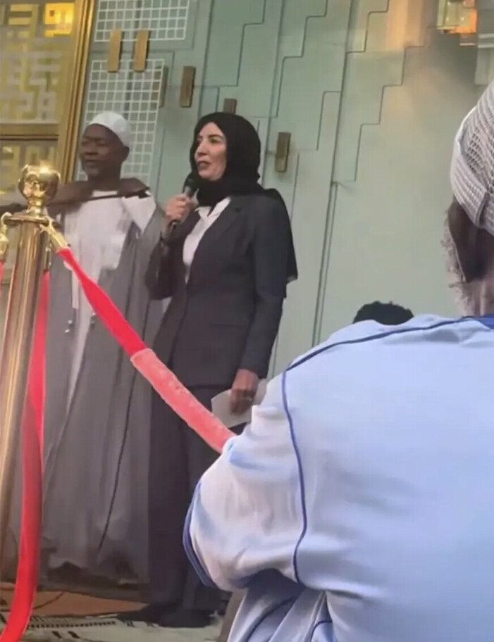 A woman in a hijab speaks into a microphone while standing next to a man in traditional attire. They are positioned near a golden railing with a red ribbon, suggesting a ceremonial or formal event. The background features a decorative architectural design, and an onlooker in a blue shirt is seated nearby.